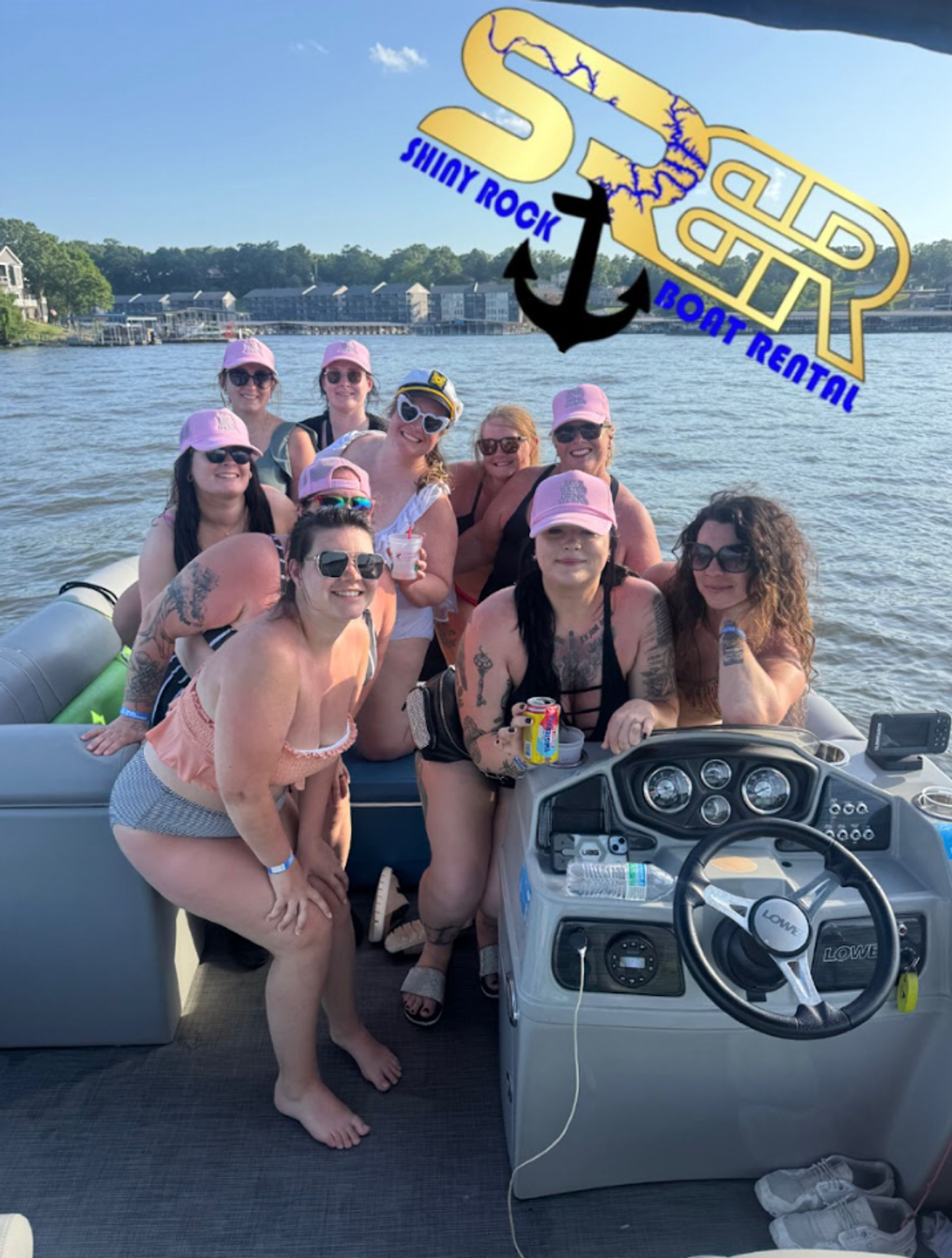 Group of women in swimsuits and matching pink caps enjoying a sunny pontoon boat party on a lake, holding drinks near the boat helm with lake houses and a tree-lined shore in the background.