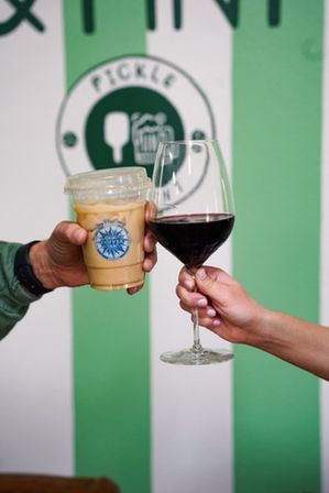 Two hands clinking drinks — a to-go iced coffee in a plastic cup and a glass of red wine against a green-and-white striped wall.