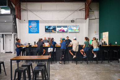 Casual patrons seated along a long wooden bar in an industrial-style brewery taproom, watching sports on three wall-mounted TVs above high stools and a concrete floor.