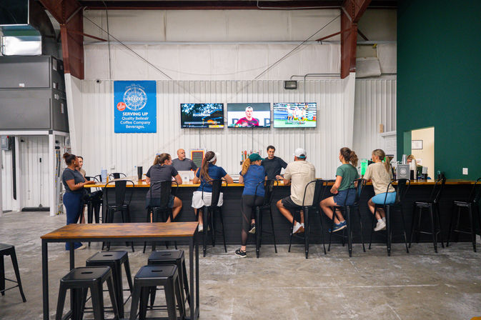 Casual patrons seated along a long wooden bar in an industrial-style brewery taproom, watching sports on three wall-mounted TVs above high stools and a concrete floor.