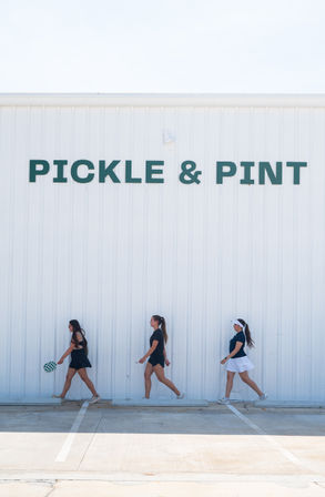 Three women in sporty outfits walk along a sunny concrete parking lot past a white corrugated storefront with bold green lettering, one carrying a checkered pickleball paddle.