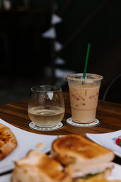 Iced coffee with green straw and a chilled water glass on coasters on a wooden café table, toasted sandwich and pizza slice blurred in the foreground — casual brunch scene.