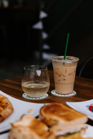 Iced coffee with green straw and a chilled water glass on coasters on a wooden café table, toasted sandwich and pizza slice blurred in the foreground — casual brunch scene.