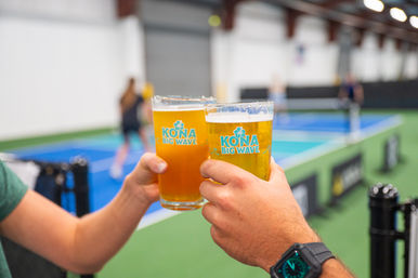 Two pint glasses of pale and amber beer clinking inside an indoor pickleball facility, with blue courts and blurred players in the background