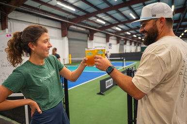 Post-game cheers: two players clink beer glasses beside an indoor pickleball court, smiling with blue courts, nets, and a sports facility interior in the background.