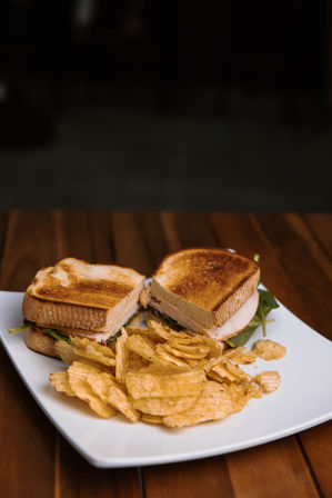 Cafe-style toasted turkey sandwich halves with fresh greens and crunchy crinkle-cut potato chips on a white plate atop a wooden table