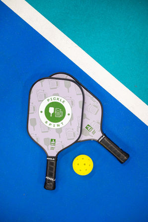 Two pickleball paddles with patterned faces and black grips beside a bright yellow pickleball on a blue court near a white diagonal line separating a teal area, playful ready-for-game composition.