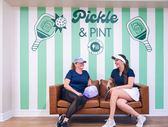 Two women in sporty outfits laugh on a brown leather couch holding pickleball paddles in an indoor pickleball lounge with a mint-green striped mural showing pickleball and pint icons.