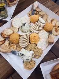 Assorted crackers and multigrain crisps with several orange cheese balls and two herbed cream-cheese spreads on a white rectangular platter with wooden-handled spreaders, set on a wooden patio table.
