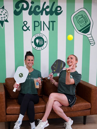 Two smiling women seated on a brown leather sofa holding pickleball paddles and an iced coffee while one tosses a bright yellow pickleball in front of a green-and-white striped, pickleball-themed wall with beer and paddle graphics.
