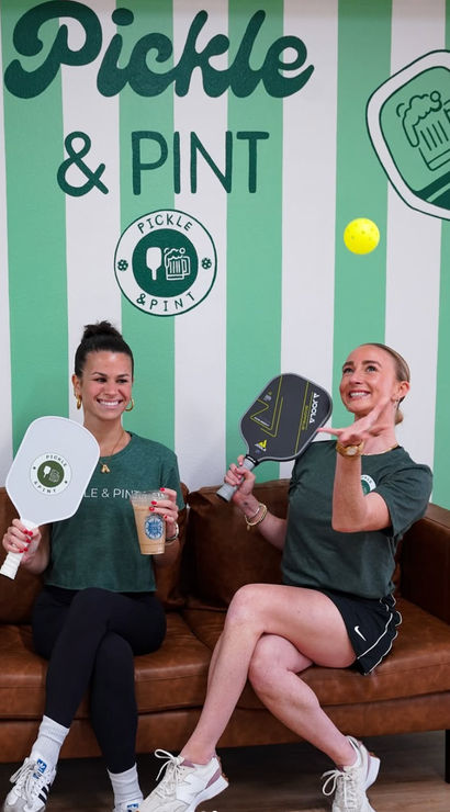 Two smiling women seated on a brown leather sofa holding pickleball paddles and an iced coffee while one tosses a bright yellow pickleball in front of a green-and-white striped, pickleball-themed wall with beer and paddle graphics.