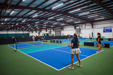 Indoor pickleball facility with multiple blue-and-green courts under steel beams, players warming up and rallying across nets beneath bright overhead lights.