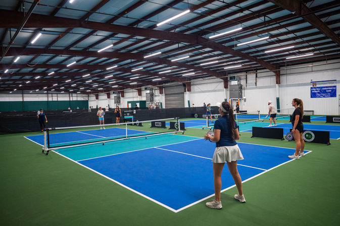 Indoor pickleball facility with multiple blue-and-green courts under steel beams, players warming up and rallying across nets beneath bright overhead lights.