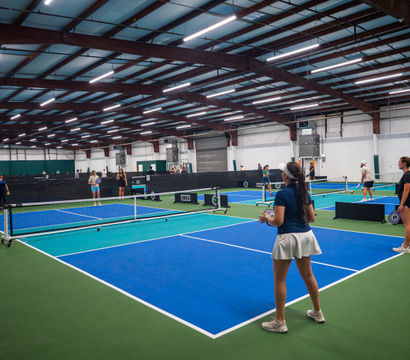 Indoor pickleball facility with multiple blue-and-green courts under steel beams, players warming up and rallying across nets beneath bright overhead lights.