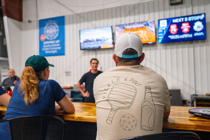Patrons seated at a wooden bar inside an indoor pickleball sports venue, man in a white cap wearing a 'Pickle & Pint' shirt with paddle, ball and bottle graphic, TV screens showing sports on the wall.
