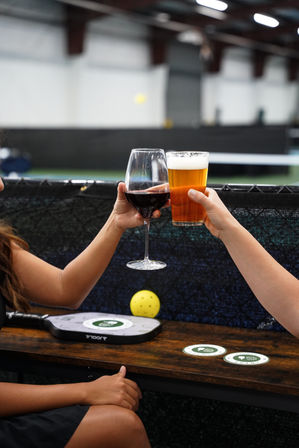 Two hands clinking a glass of red wine and a pint of beer over a wooden table beside a pickleball paddle and yellow pickleball in an indoor court — casual cheers scene.
