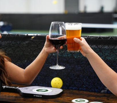 Two hands clinking a glass of red wine and a pint of beer over a wooden table beside a pickleball paddle and yellow pickleball in an indoor court — casual cheers scene.