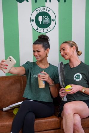 Two women in green shirts take a selfie on a brown leather couch in a pickleball-themed café, holding an iced coffee, a paddle and a neon-yellow pickleball against a green-and-white striped wall.