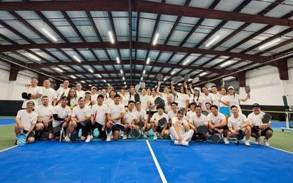Large group of recreational pickleball players in white shirts smiling and posing with paddles for a group photo on blue indoor courts under a high metal roof