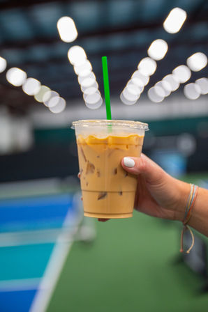 Hand with white nail polish and colorful bracelets holding an iced coffee in a clear plastic cup with green straw against a blurred indoor sports court and bokeh overhead lights.