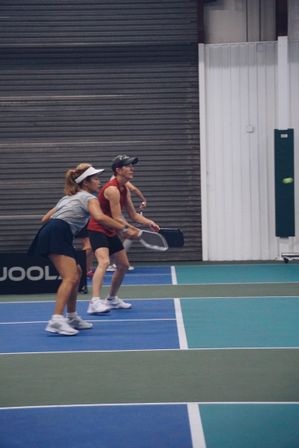 Two players on an indoor pickleball court, crouched and ready to return a shot—woman in a visor and skirt and partner in a red sleeveless top holding paddles on a blue-green court with the ball in play.