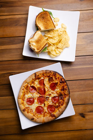 Top-down: pepperoni pizza and a toasted sandwich with potato chips on white square plates over a rustic wooden table.