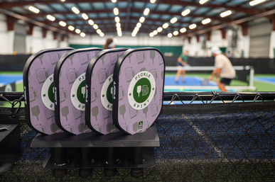 Stacked pickleball paddles on a court-side fence at an indoor pickleball facility with players rallying on blue courts under bright ceiling lights.