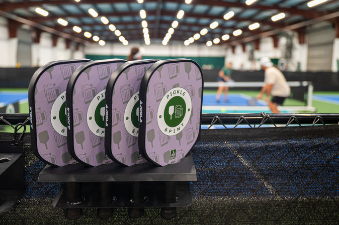Stacked pickleball paddles on a court-side fence at an indoor pickleball facility with players rallying on blue courts under bright ceiling lights.