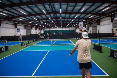 Player in a white cap prepares to serve on bright blue indoor pickleball courts with multiple nets, other players and a high warehouse-style ceiling