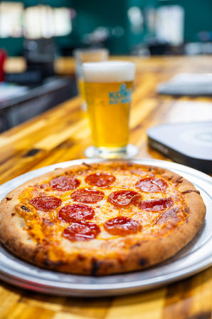 Cheesy pepperoni pizza on a round metal tray atop a wooden bar counter with a frothy pint of craft beer blurred in the background — casual pizzeria scene