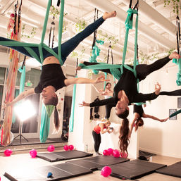 Group aerial yoga class in a bright indoor studio: participants suspended upside-down in teal silk hammocks over mats, with pink balloons and hanging greenery adding playful decor.