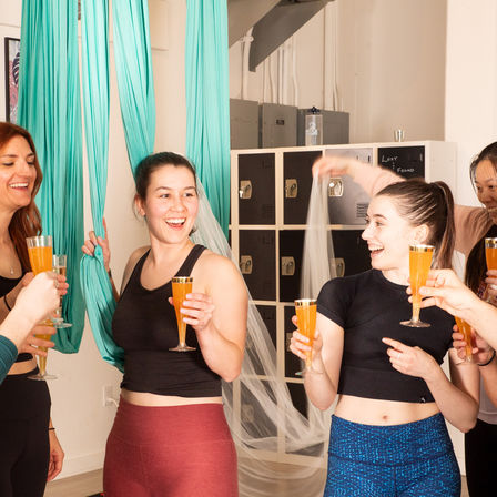 Group of women in workout clothes toasting with mimosas in an aerial yoga studio under teal silk hammocks, celebrating a bachelorette party.