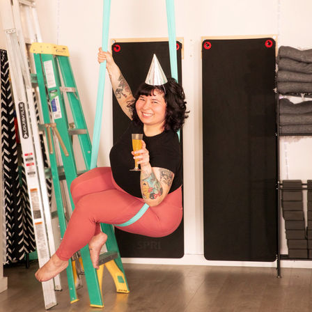 Smiling person in a party hat suspended in a turquoise aerial yoga hammock, holding a drink in an indoor yoga studio with ladders, mats and stacked props.