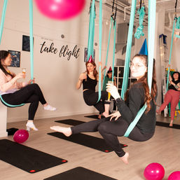 Group aerial yoga class in a bright indoor studio — participants suspended in teal silk hammocks wearing party hats and holding drinks, with yoga mats, pink balloons and 'take flight' wall art.