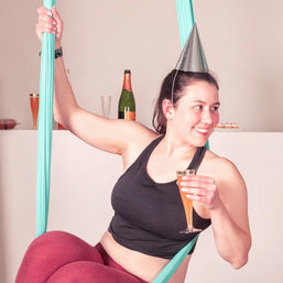 Celebratory aerial yoga scene: person in a metallic party cone hat smiling and sipping champagne while seated in a turquoise aerial hammock, with a champagne bottle and cupcakes on a counter behind them.