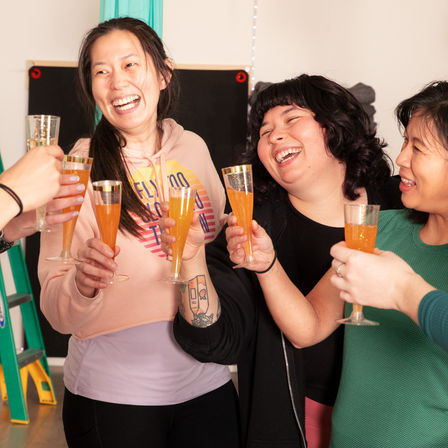 Group of friends laughing and clinking tall flutes of orange sparkling cocktails at a cozy indoor gathering, casual clothes, a visible forearm tattoo and a ladder in the background.