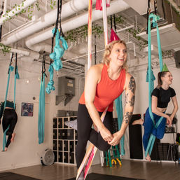 Two women enjoying an indoor aerial yoga class on teal silks — woman in an orange tank with a floral tattoo wearing a pink party hat, bright modern fitness studio