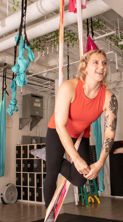 Two women enjoying an indoor aerial yoga class on teal silks — woman in an orange tank with a floral tattoo wearing a pink party hat, bright modern fitness studio