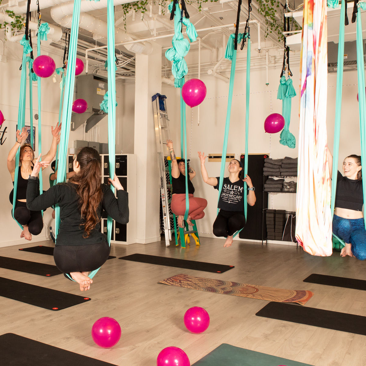 Playful aerial yoga class in a bright indoor studio with participants suspended in teal hammocks above mats, colorful pink balloons scattered on the floor and floating around.