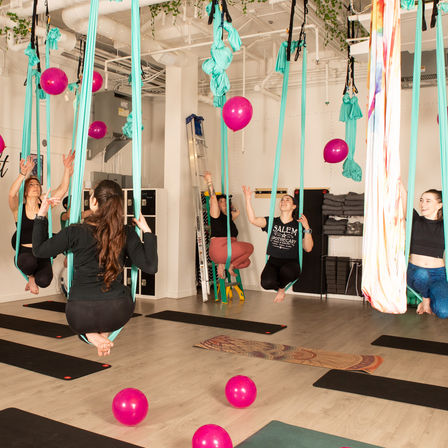 Playful aerial yoga class in a bright indoor studio with participants suspended in teal hammocks above mats, colorful pink balloons scattered on the floor and floating around.