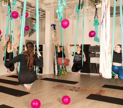 Playful aerial yoga class in a bright indoor studio with participants suspended in teal hammocks above mats, colorful pink balloons scattered on the floor and floating around.