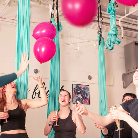 Group celebrating in an indoor aerial yoga studio with turquoise silks, bright pink balloons floating overhead, and smiling participants holding champagne flutes.