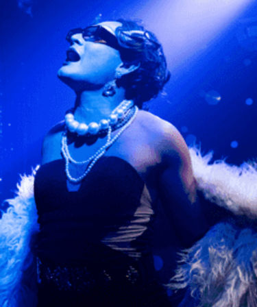 Drag performer in vintage glam — strapless black dress, layered pearl necklaces and feather boa, singing under blue spotlight on a cabaret/nightclub stage.