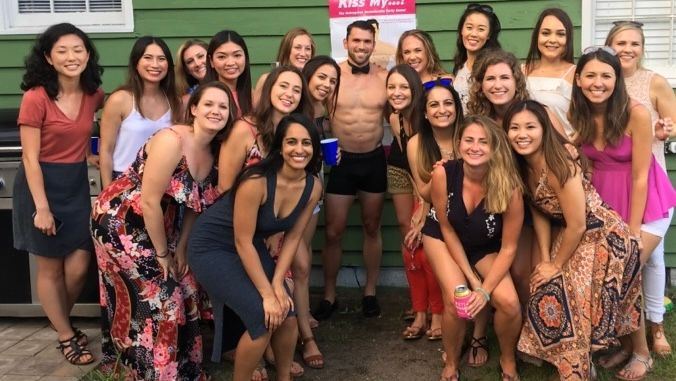 Smiling group of women at a backyard summer party posing around a shirtless male entertainer in black shorts and bow tie, colorful summer dresses, drinks and green house siding in the background.