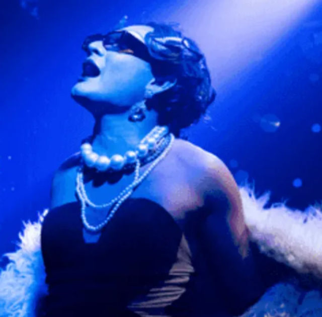 Live cabaret performer under blue spotlight wearing sunglasses, layered pearl necklaces, strapless dress and feather boa, head tilted back and mouth open mid-song on stage.