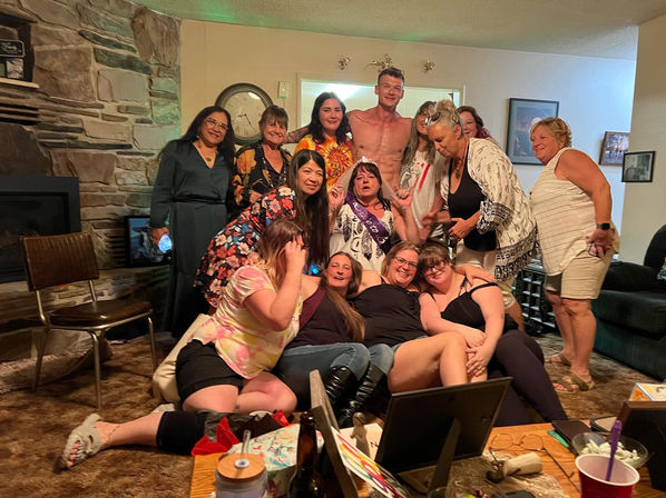 Around a dozen adults gathered in a cozy living room for a party, a woman wearing a sash and tiara at center, friends lounging on a couch and floor, stone fireplace and coffee table with drinks visible.