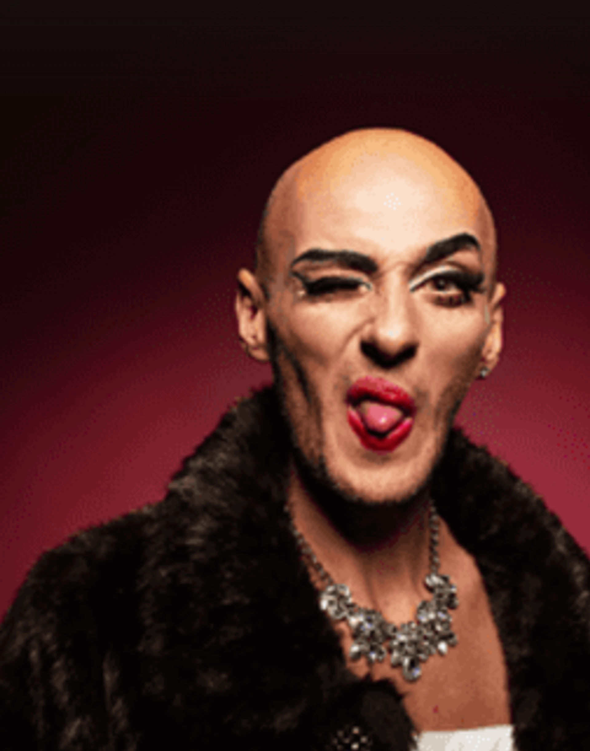 Bald drag performer portrait with bold eye makeup and red lipstick sticking out tongue, wearing a faux-fur coat and statement necklace against a deep maroon studio background.