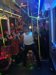 Party bus interior with neon lights: a male dancer in a white tank gives a playful lap dance to a seated woman while a crowd of women wearing matching pink flower crowns cheer, laugh, and film on their phones near yellow handrails.