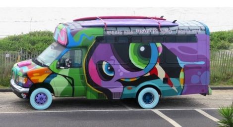 Vibrant graffiti-covered shuttle van with a large stylized eye, teal and purple wheels, and abstract colorful shapes parked on a coastal road by sand dunes and a wooden fence.
