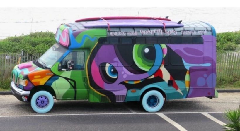 Vibrant graffiti-covered shuttle van with a large stylized eye, teal and purple wheels, and abstract colorful shapes parked on a coastal road by sand dunes and a wooden fence.